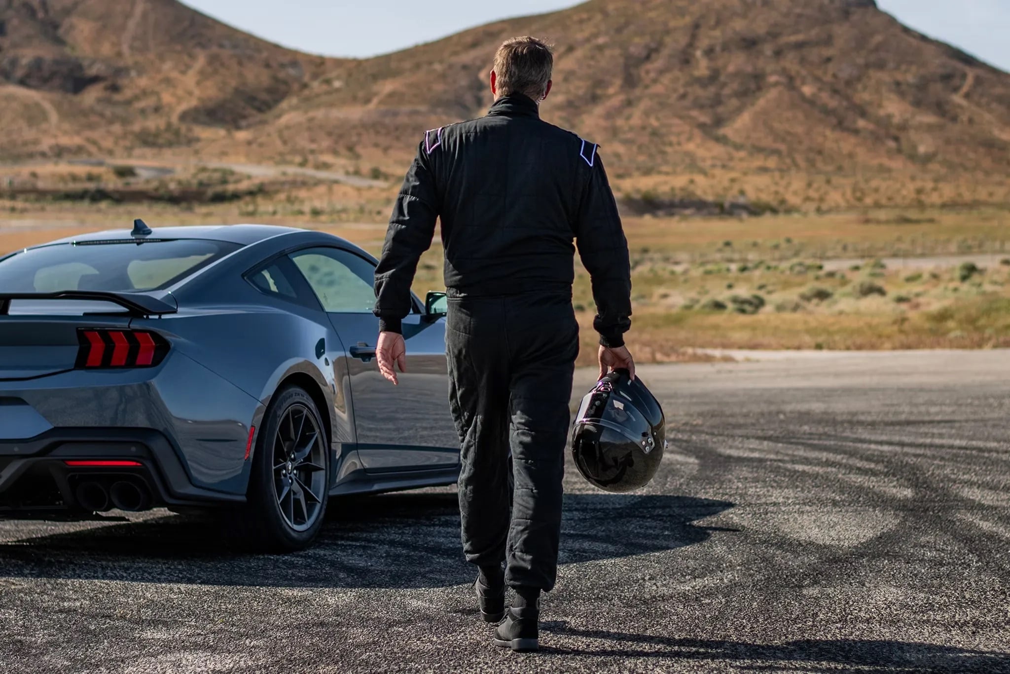 Driver in racing suit walking toward a 2026 Ford Mustang Dark Horse™ on a racetrack, holding a black helmet with desert hills in the background.
