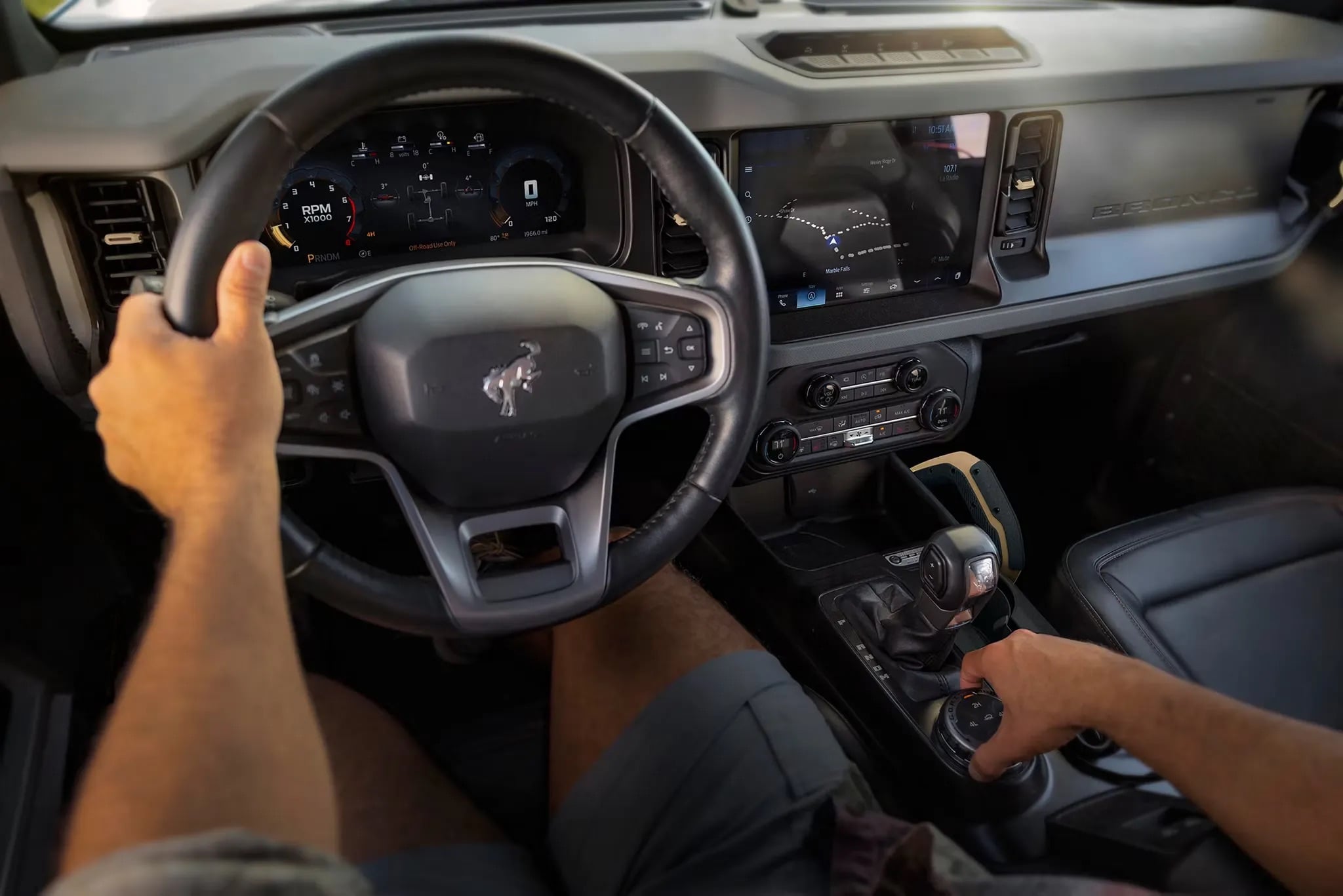 Driver’s point of view inside a Ford Bronco, showing the steering wheel, digital instrument cluster, large centre touchscreen with SYNC® 4, and a clear view of the trail ahead through the upright windshield.