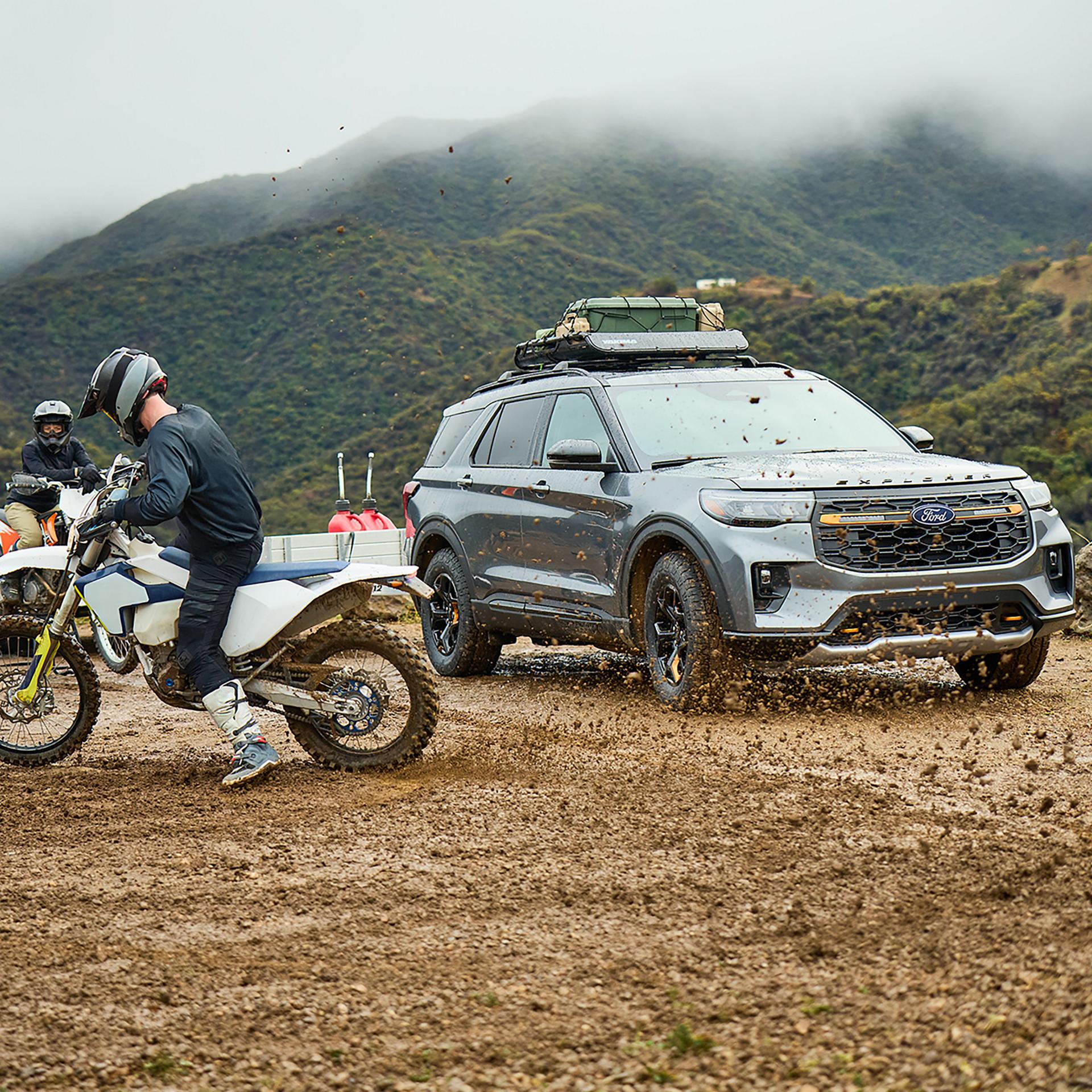 Ford Explorer parked beside a dirt bike and rider in a mountain clearing