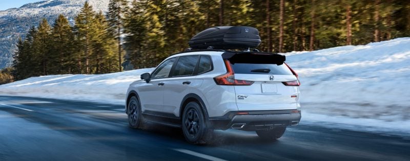 White Honda CR-V with a rooftop cargo box driving on a winter road beside snowbanks and evergreen trees.