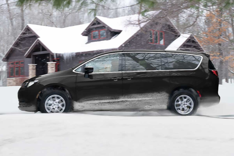 2026 Chrysler Grand Caravan in black driving through snow near a cozy cabin, showcasing confident winter handling