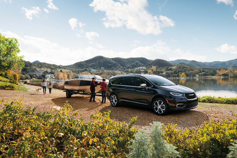2026 Chrysler Pacifica in black towing a boat beside a scenic lake with rolling hills in the background.