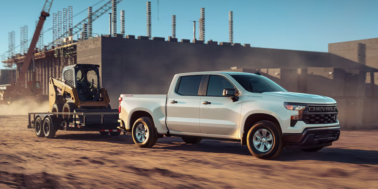 Three quarters view of a white 2025 Chevrolet Silverado LD Pickup Truck towing a skid steer loader on a construction site.