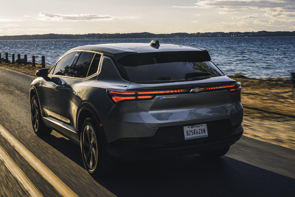 Rear view of the 2026 Chevrolet Equinox EV driving along a lakeside road at sunset, emphasizing its modern taillight design