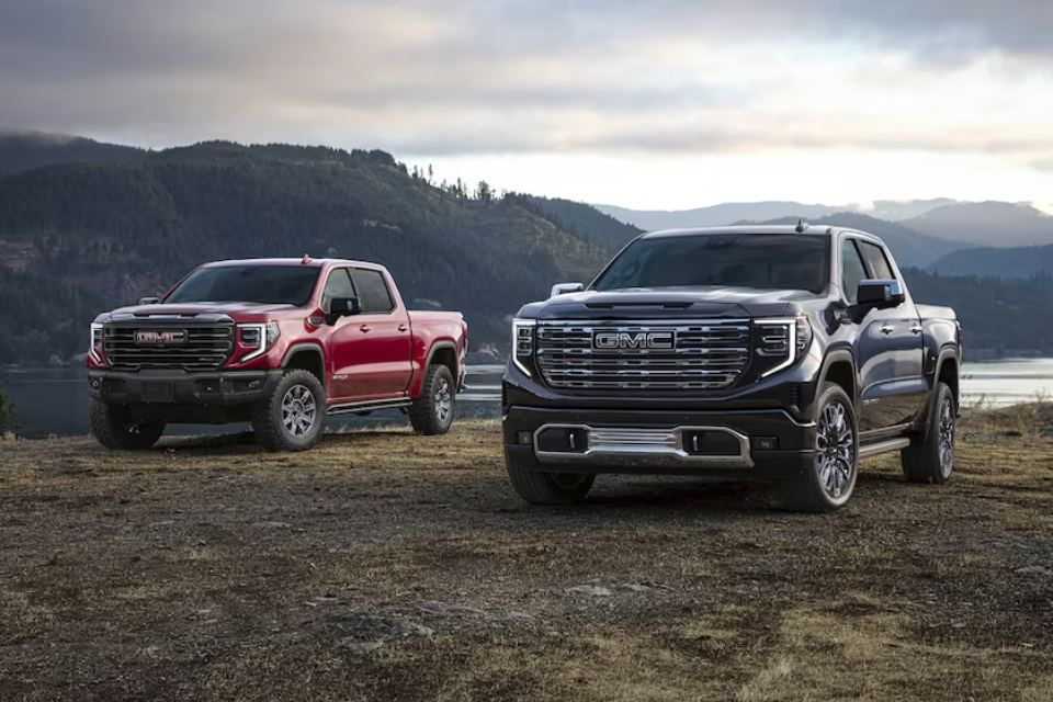 Two 2026 GMC Sierra 1500 trucks, one red and one black, parked in a scenic mountain area, showcasing rugged strength and bold design