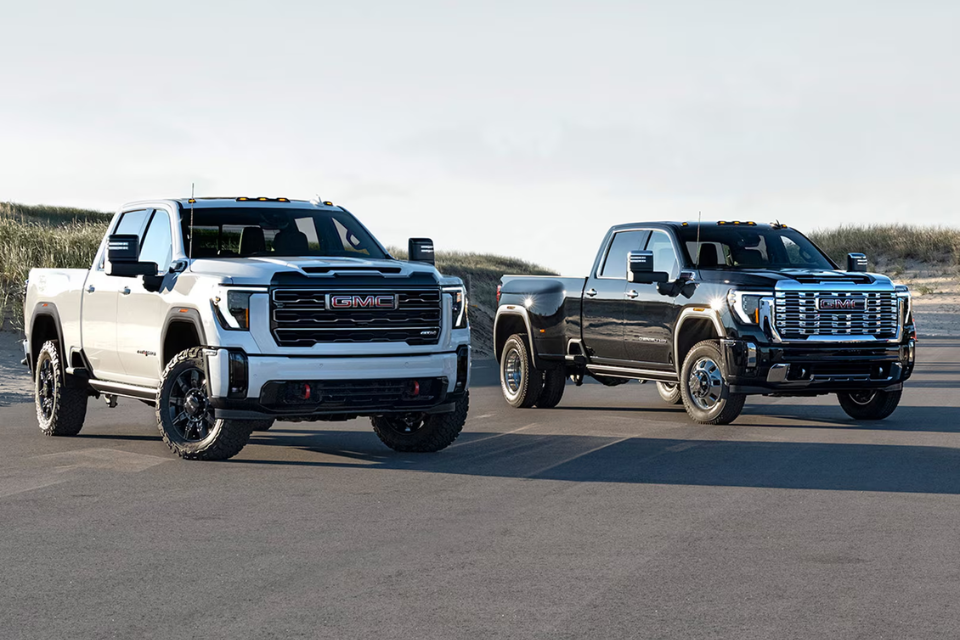2026 GMC Sierra HD trucks parked side by side on a coastal road, showcasing the bold front-end design of the AT4 and Denali trims