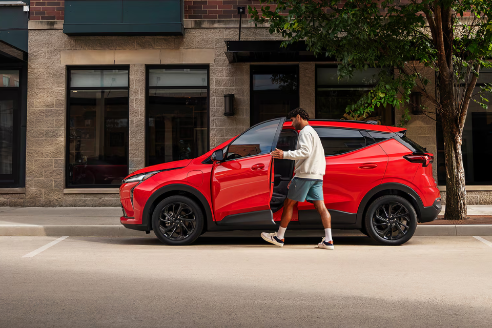 Side view of the 2027 Chevrolet Bolt EV in red with driver entering the vehicle on a city street