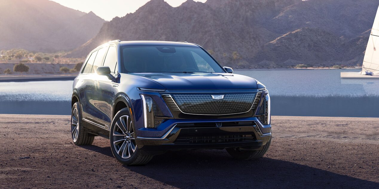 Parked near a body of water with mountains in the background, featuring illuminated grille and signature Cadillac styling