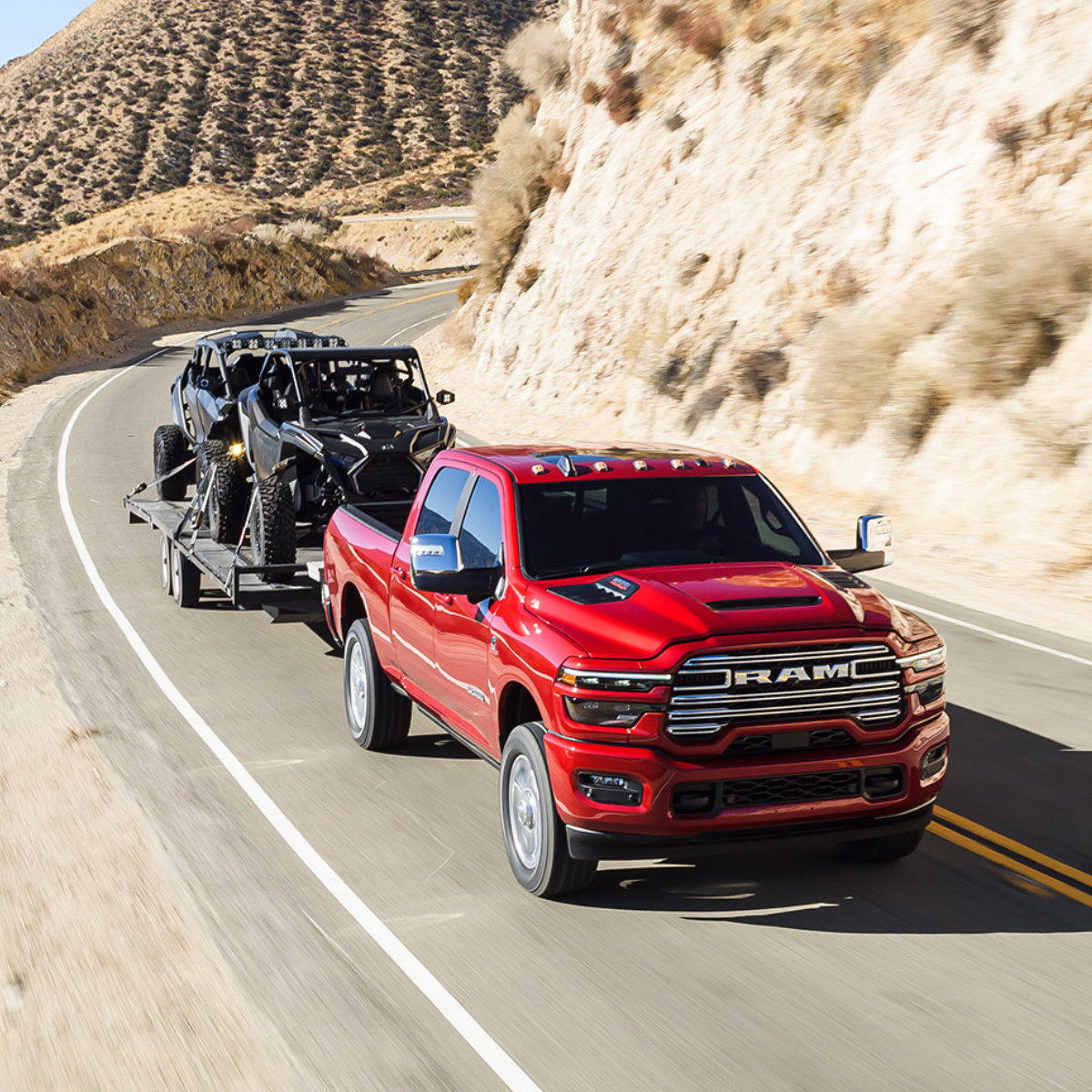 Red 2025 Ram 2500 HD towing two side-by-side off-road vehicles on a trailer along a winding mountain highway