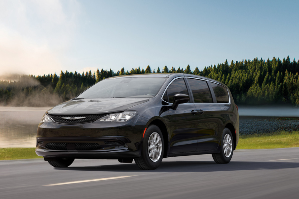 2026 Chrysler Grand Caravan driving along a lakeside road surrounded by trees, highlighting its refined exterior styling